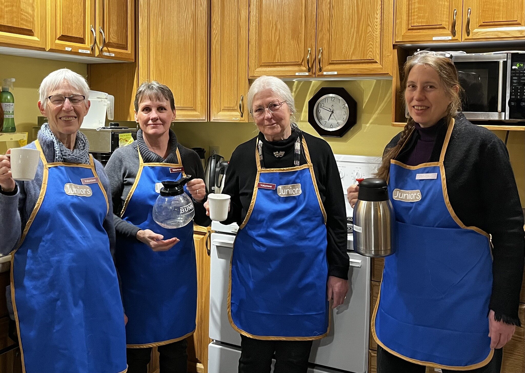 Four Shuswap Theatre volunteers in aprons smiling together while serving refreshments.